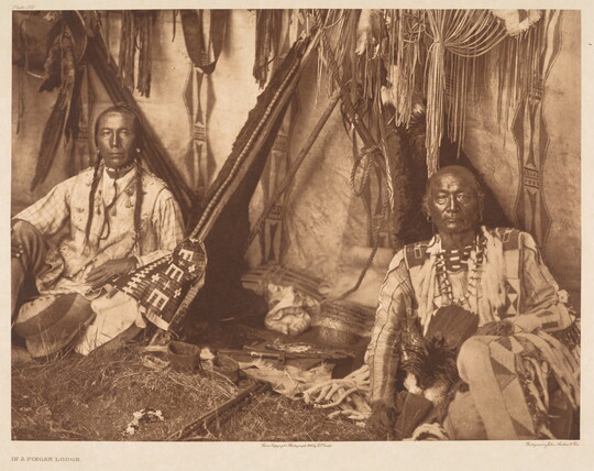 A sepia-toned photograph of two Indigenous adults sitting inside a Native American lodge wearing cultural dress.