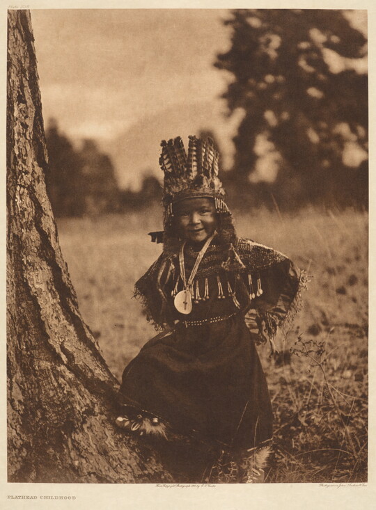 A sepia-toned portrait photograph of a Native American child in traditional dress and headdress, standing next to a tree with hands on hips.