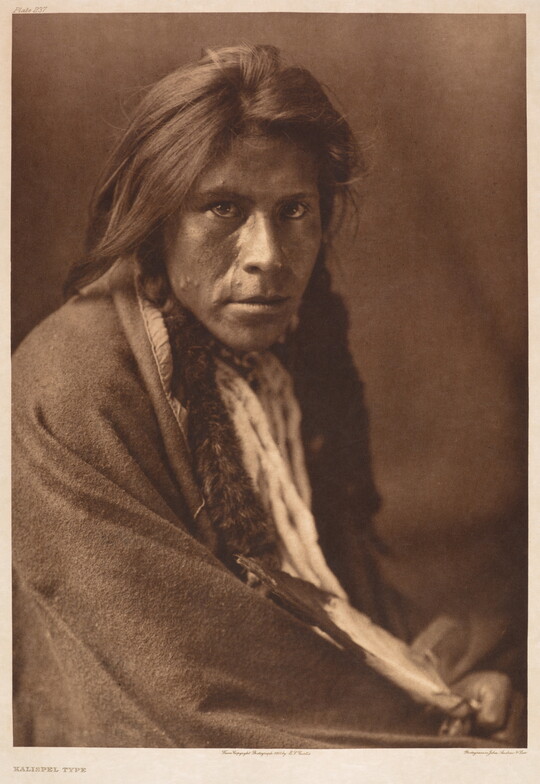 A sepia-toned portrait photograph of an Indigenous man with a blanket around his shoulders leaning toward the camera.