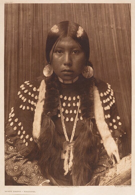 A sepia-toned portrait photograph of an Indigenous girl from the waist up dressed in traditional clothing.
