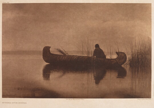 A sepia-toned photograph of an adult figure sitting in a canoe on calm water.