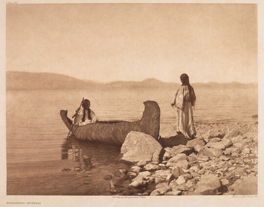 A sepia-toned photograph of a Native American person sitting in a canoe on the water as another person stands on the rocky shore.