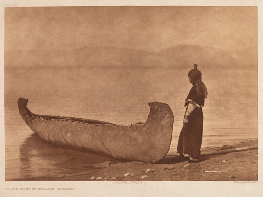 A sepia-toned photograph of a canoe on the edge of the shore and an Indigenous person standing beside it.