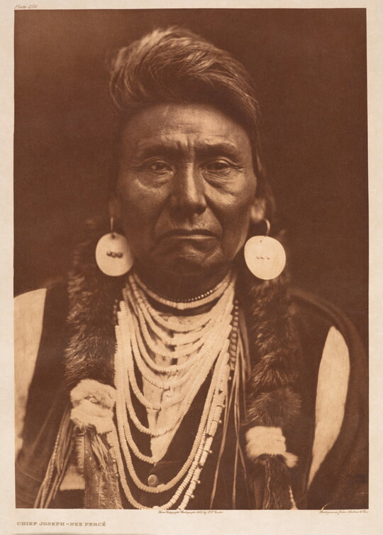 A sepia-toned portrait photograph of an Indigenous man wearing cultural jewelry and clothing.
