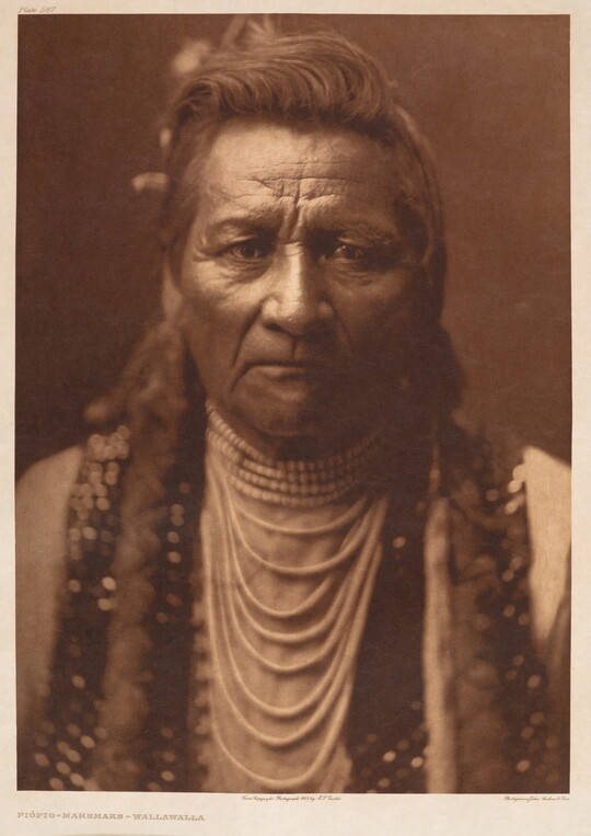 A sepia-toned portrait photograph of a Native American man from the chest up in traditional dress, looking at the viewer.