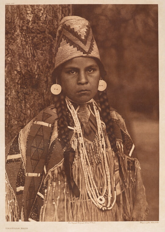 A sepia-toned portrait photograph of a Native American girl with braids, wearing a tall decorative hat and traditional clothing.