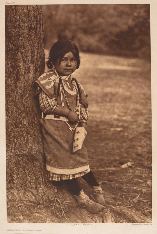 A sepia-toned portrait photograph of a young Native American girl in traditional dress leaning against a tree.