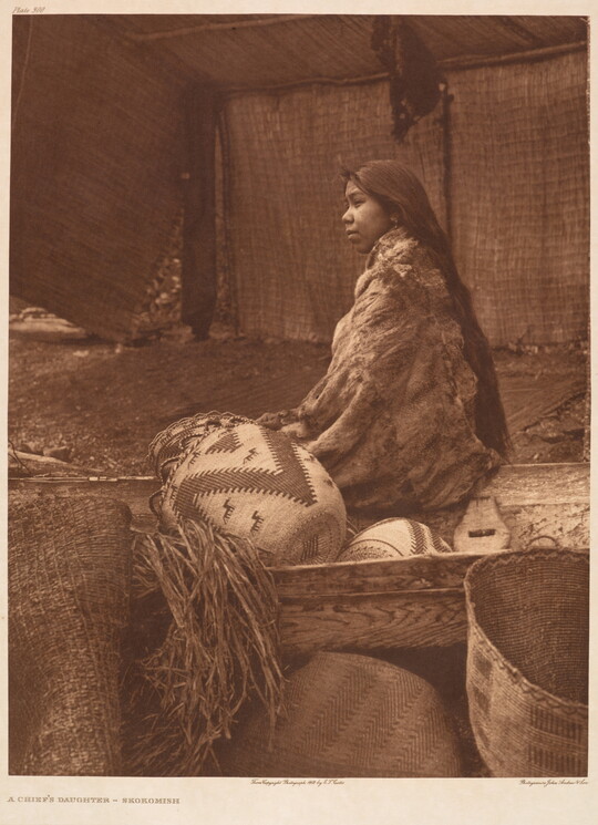 A sepia-toned photograph of a Native American woman wrapped in a blanket, sitting with back to the viewer next to a wood crate containing woven baskets.