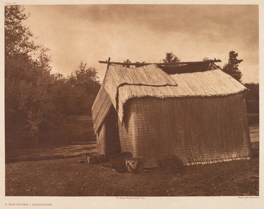 A sepia-toned photograph of a wood structure covered with woven straw mats and a few pots at the entrance.