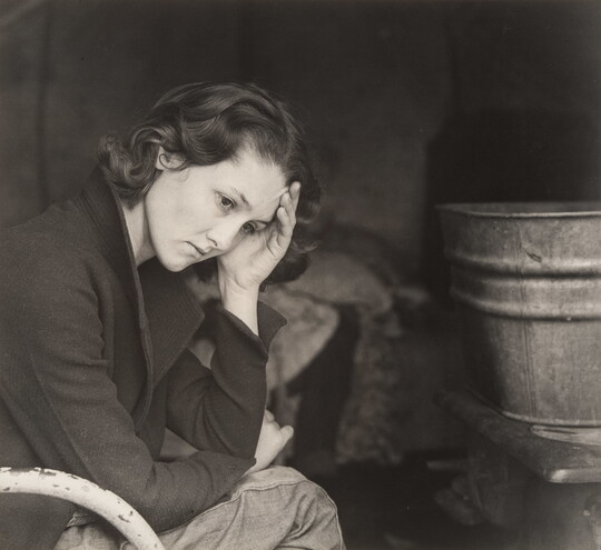 A black-and-white portrait photograph of a young, White woman sitting in a chair with her head in her hand.