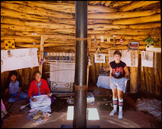 A color photograph of Native American people with a loom and crafts inside a building made of stripped logs.