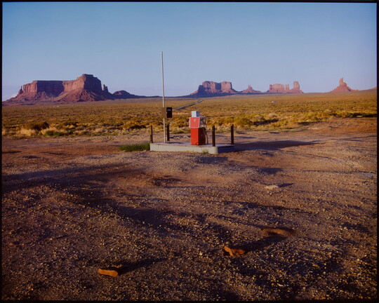A color photograph of a lone gas pump on a concrete pad in a desert landscape with mesas and buttes in the background.