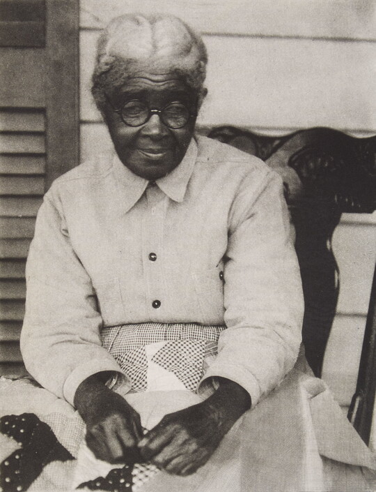 A black-and-white photograph of a seated, elderly Black woman holding a quilt on her lap.