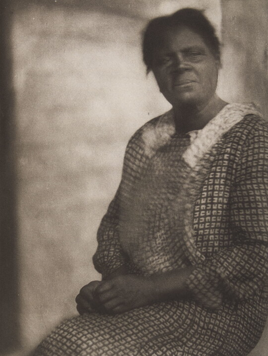 A black-and-white, unfocused photograph of a seated Black woman in a patterned dress, gazing out.