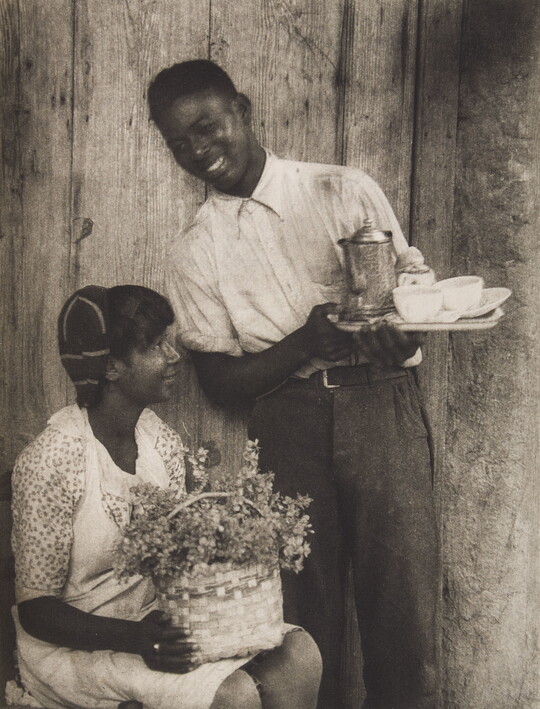 A black-and-white photograph of a young Black woman holding a basket of flowers on her lap and a young Black man standing next to her with a coffee service, both smiling.