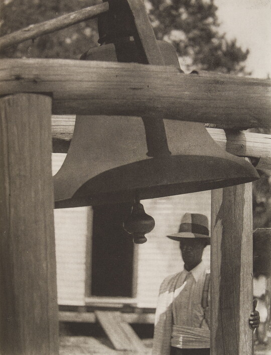 A black-and-white photograph of a Black man in a hat standing to the side of a wooden structure that holds a large bell.
