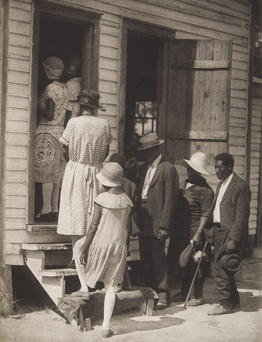 A black-and-white photograph of a group of Black people dressed nicely going up steps into a clapboard building.