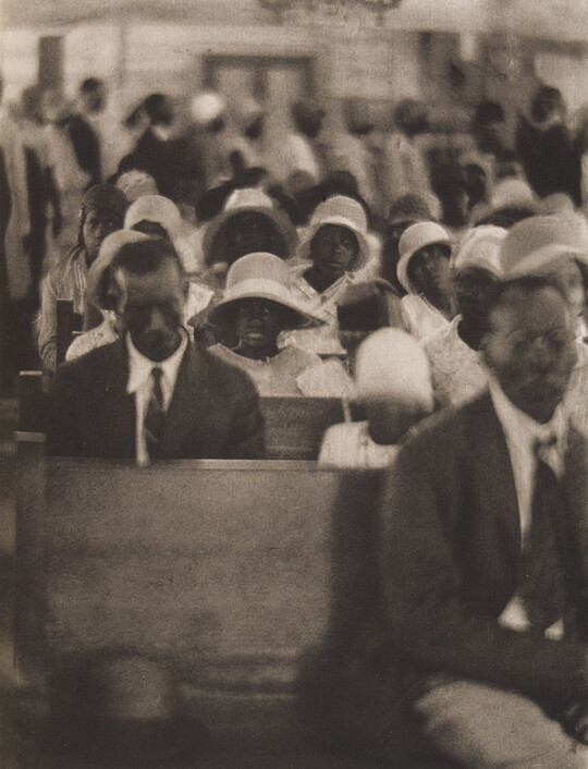 A soft-focus, black-and-white photograph of Black congregants seated in crowded church pews and standing in the back.