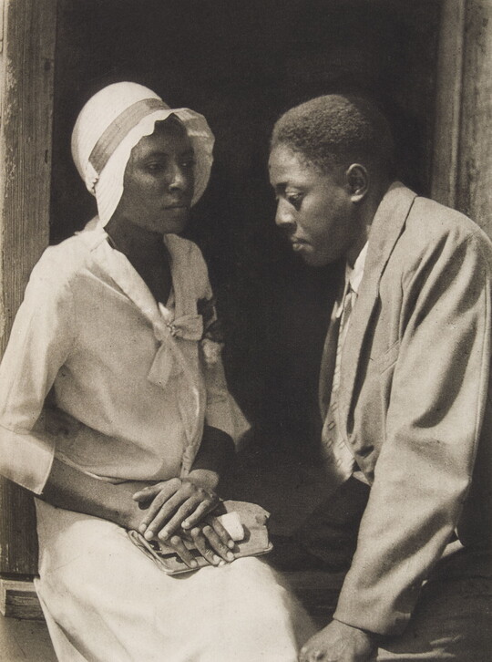 A black-and-white photograph of a young Black couple dressed nicely and seated in a doorway.
