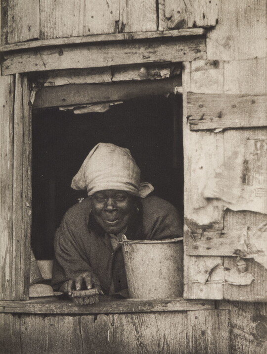 A black-and-white photograph of a Black woman wearing a bandana over her hair, holding a scrub brush and leaning over a pail from the window of a wood building.