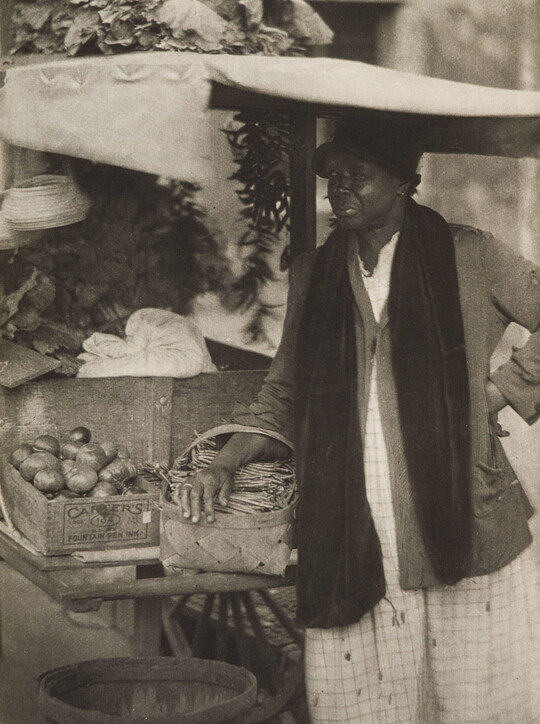 A black-and-white photograph of a Black woman standing with a basket on her arm next to a cart of crated vegetables.