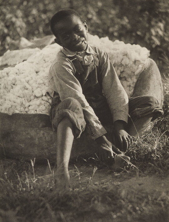A black-and-white photograph of a smiling young Black boy sitting on a bag of cotton.
