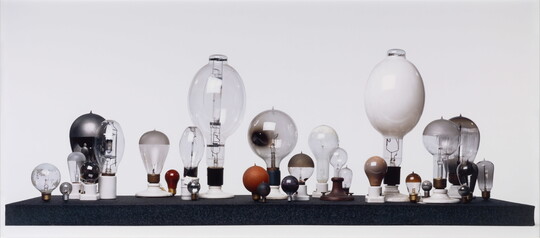 A color photograph of lightbulbs of many shapes and sizes, arranged with their socket sides down on a black platform in front of a white background.
