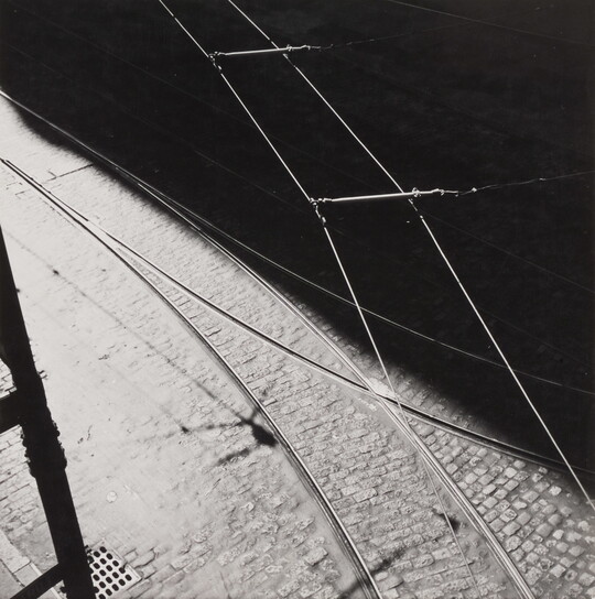 A black-and-white abstract photograph of tram tracks, a brick street, and wires divided diagonally by a hard shadow.