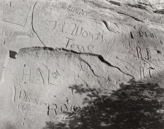 A black-and-white, tightly focused photograph of a large rock with carved inscriptions, including various initials and the words "Ft. Worth, Texas."