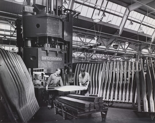 A black-and-white photograph of two men in a factory holding an airplane propeller surrounded by racks of propellers.