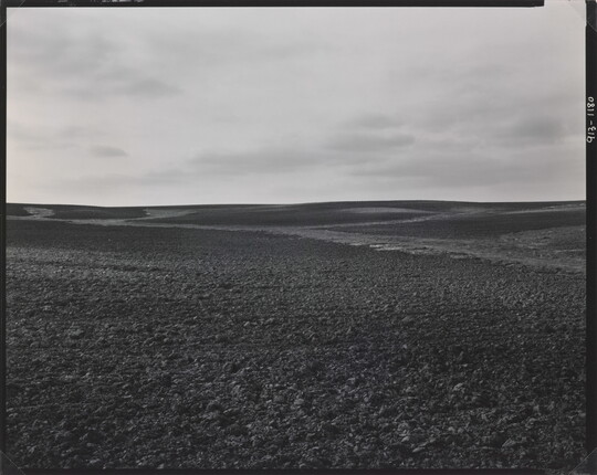 A black-and-white photograph of a dirt landscape with subtle hills under a cloudy sky.