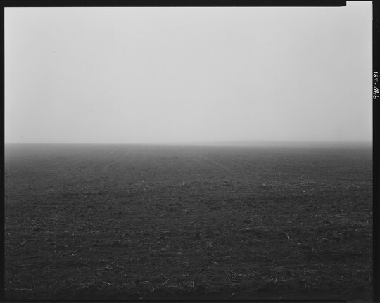 A black-and-white photograph of a dirt landscape with a hazy horizon and a subtle hill in the distance.