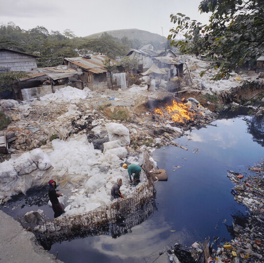 A color photograph of people sorting through a garbage dump next to water, several shacks in the background, and a fire going next to the water.