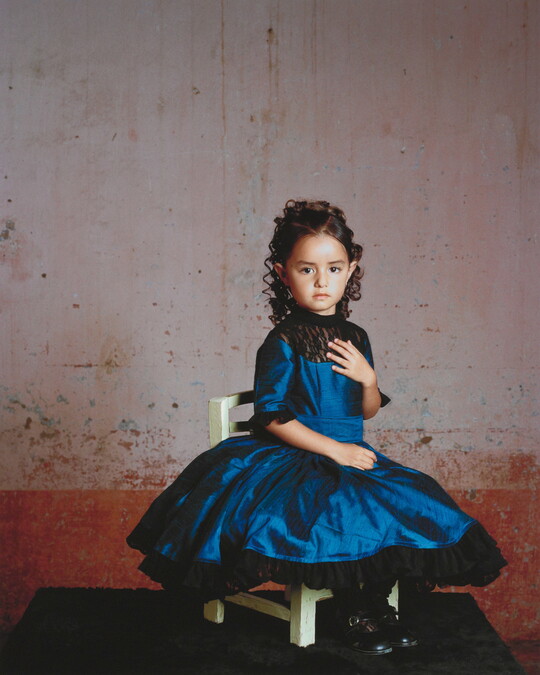A color photograph of a young girl in a formal blue dress posed on a white chair.