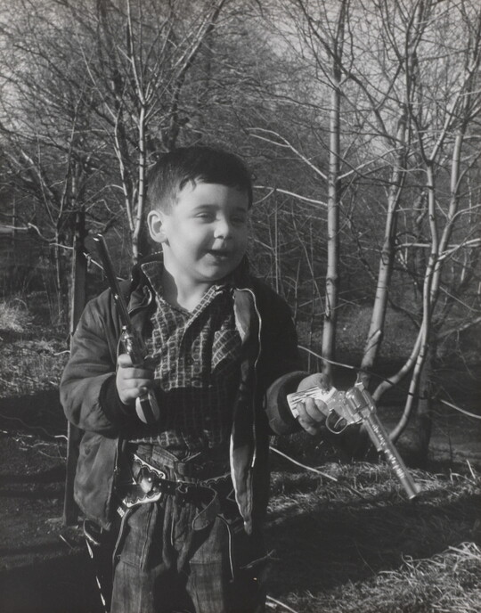 A black-and-white photograph of a White boy playing with two toy revolvers in a wooded setting.