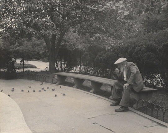 A black-and-white photograph of an older White man with a beard sitting on a stone bench in a park.