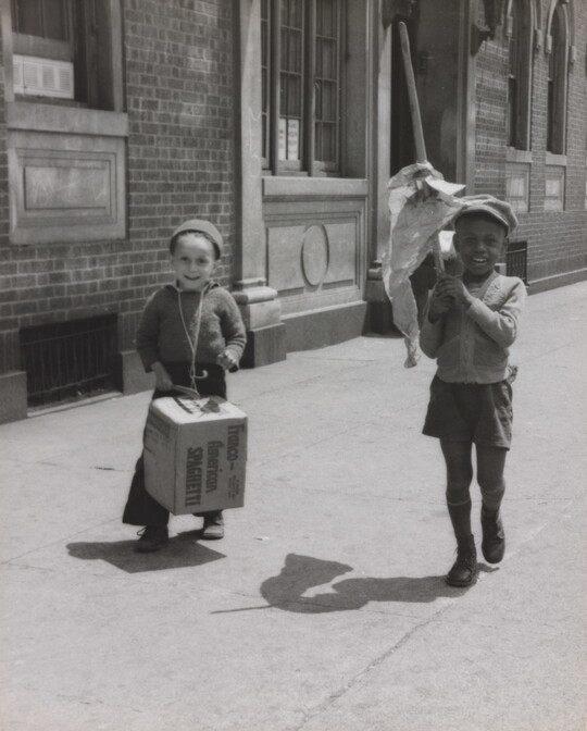 A black-and-white photograph of a White child banging a box like a drum and a Black child waving a flag of crumpled paper as they march on a city sidewalk.