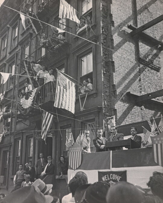 A black-and-white photograph of a man making a speech on a platform with people watching and American flags hanging from apartment windows above.