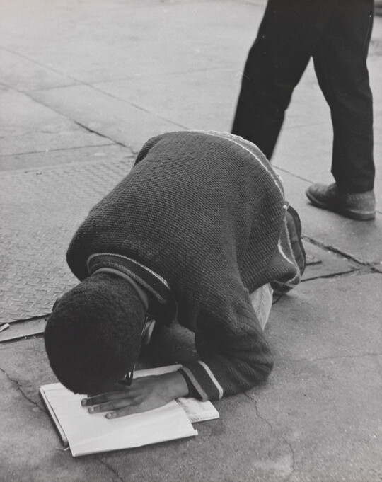 A black-and-white photograph of a Black boy hunched over his knees on pavement, his hand on a book and face close to his hand.