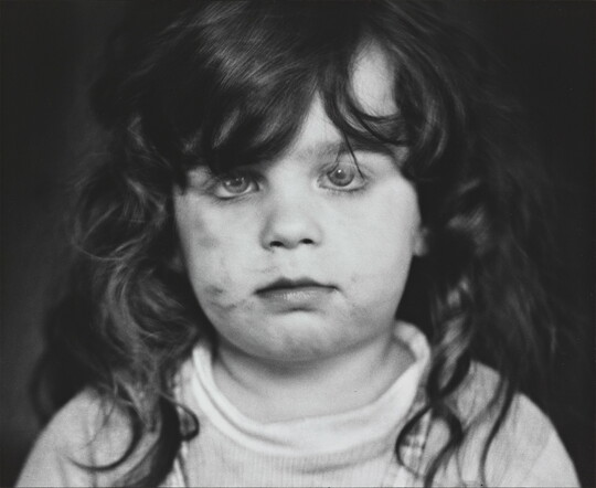 A black-and-white photograph of a White, toddler girl with dark, curly hair and dirt on her cheeks looking directly at the viewer.