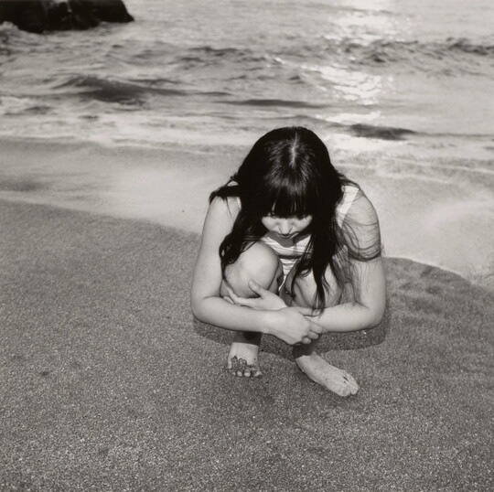 A black-and-white photograph of a young White girl with long dark hair crouched on a shoreline looking at the sand.