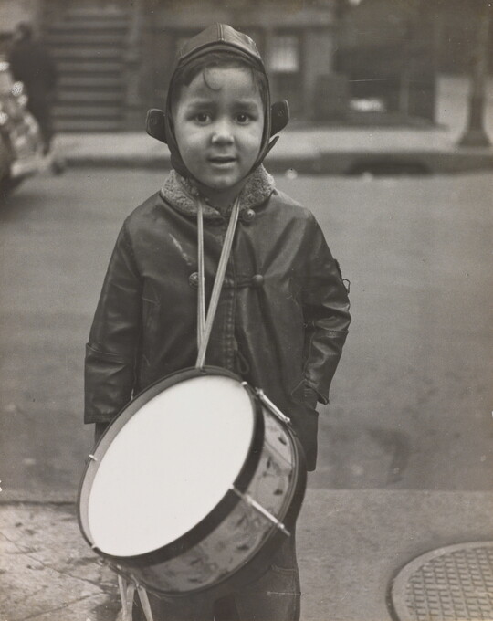 A black-and-white photograph of a warmly dressed young boy with medium-toned skin wearing a drum from his neck and looking directly into camera.