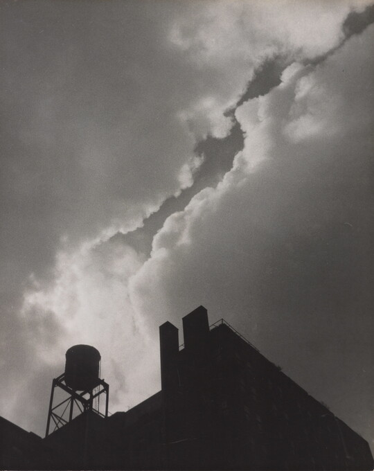 A black-and-white photograph taken from the ground looking up of silhouettes of industrial buildings and a water tower against a cloudy sky.
