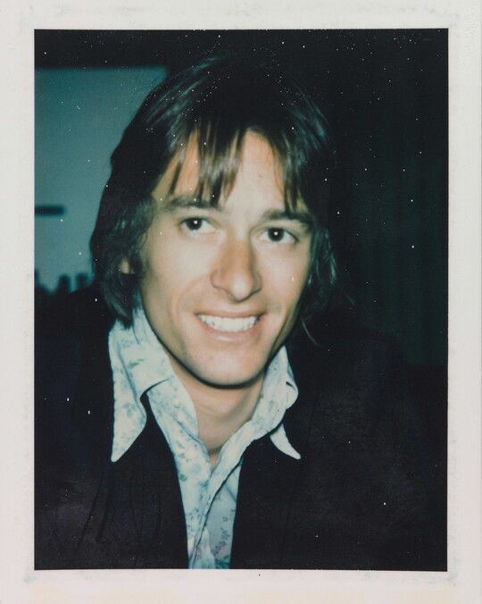An informal color photograph of a smiling White man from the shoulders up with longish hair, wearing an open collar shirt and dark jacket.