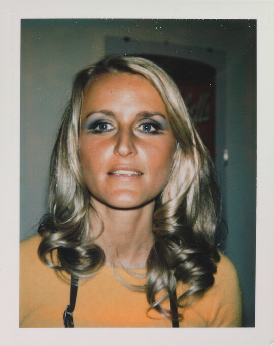 An informal color photograph of a White woman from the shoulders up, with blonde hair, blue eyeshadow, and a tanned face.