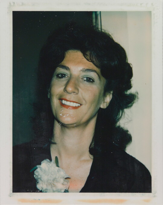 An informal color photograph of a smiling White woman from the shoulders up with dark, curly hair, red lipstick, and a white carnation on her black shirt.