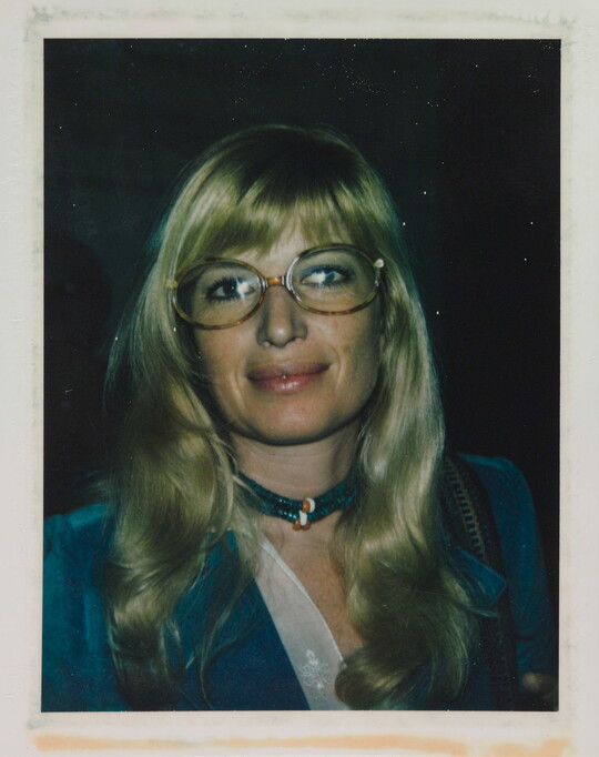 An informal color photograph of a White woman with blonde hair and tortoise-shell glasses wearing a blue suit and necklace.