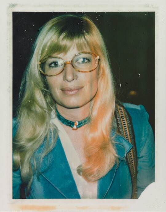 An informal color photograph of a White woman with blonde hair and tortoise-shell glasses wearing a blue suit and necklace.