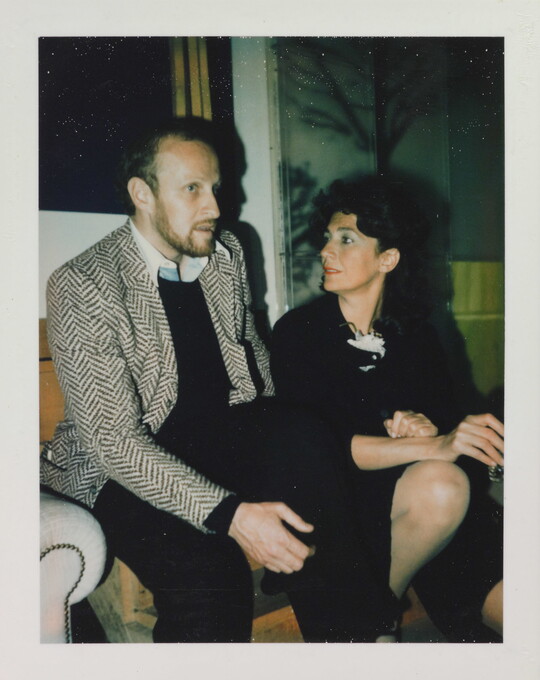 A color photograph of a White man and woman seated on a couch with the woman gazing in the man's direction as he seems to speak.