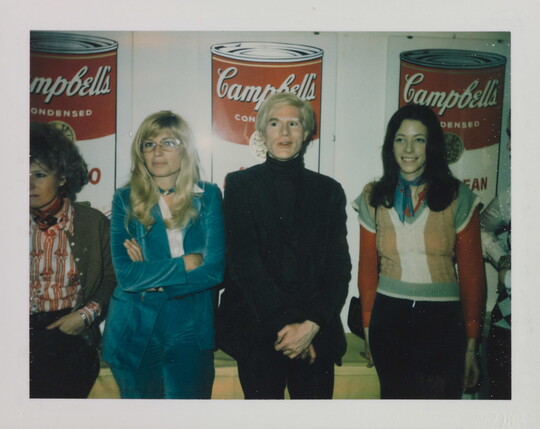A color photograph of four White people standing in front of three iconic images of "Campbell's Soup" cans.